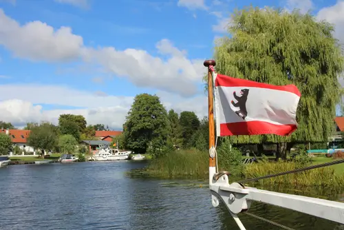 Ein Kanal, blauer Himmel, im Hintergrund sind Häuser und Boote zu sehen, im Vordergrund die Flagge des Landes Berlin am Boot angebracht