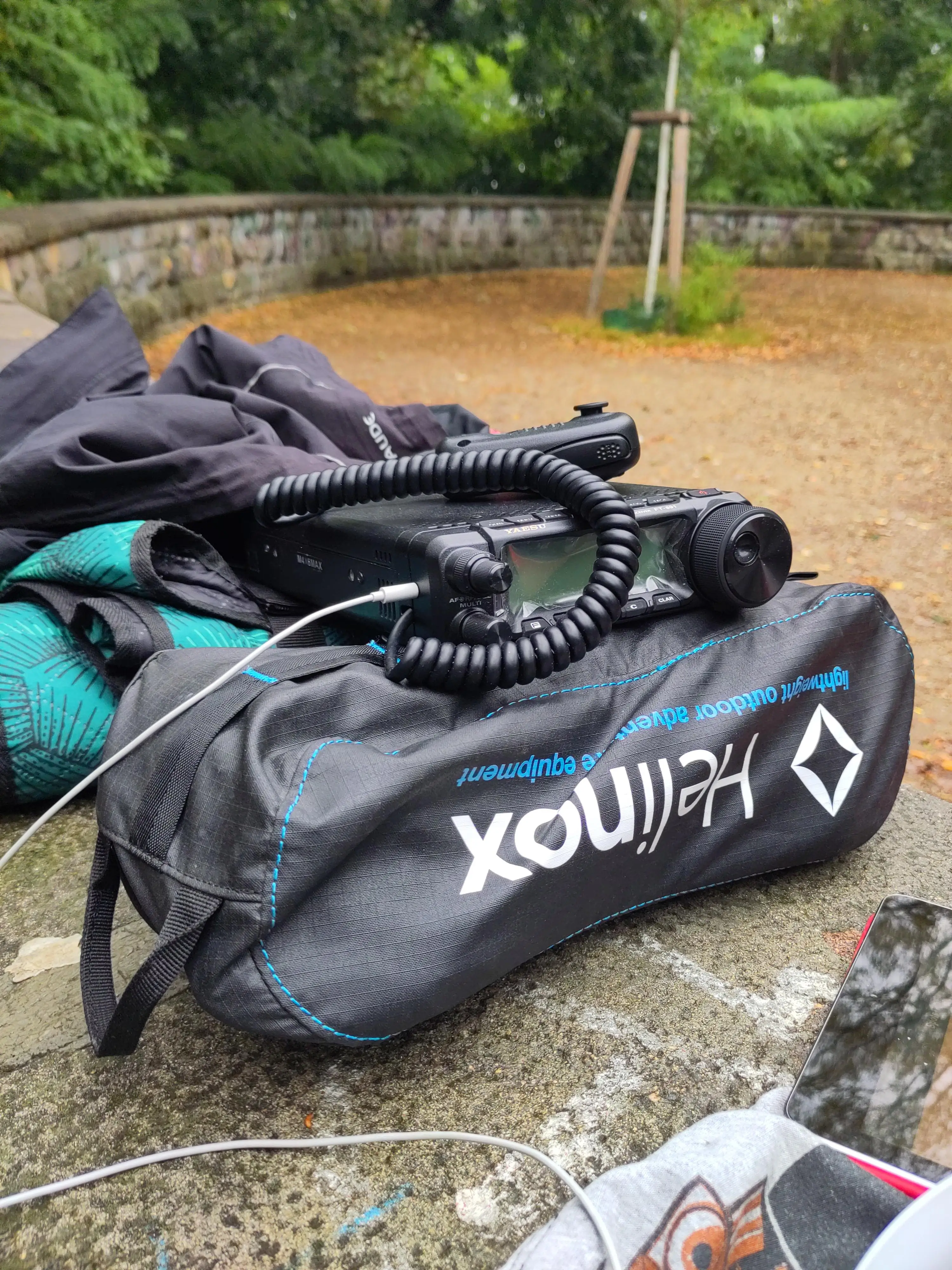 Auf einem steinernen Untergrund steht ein Funkgerät aufgebaut auf der Hülle eines Klappstuhls und einer Picknickdecke. Im Hintergrund sieht man die Andeutung des Rundells auf dem großen Bunkerberg im Volkspark Friedrichshain